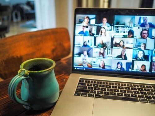 A home environment with laptop showing Zoom chat with several participants and a mug near the computer.